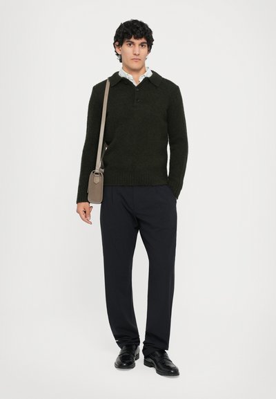 Dunkelgrüner Wollpullover mit Kragen und einer Knopfleiste mit drei Knöpfen, kombiniert mit locker sitzenden schwarzen Hosen und schwarzen Schuhen. Tanfarbige Schultertasche.
