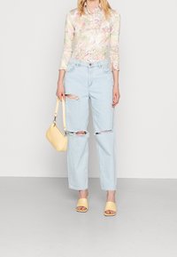 Mujer vestida con jeans rasgados de color azul claro, camisa de manga larga con estampado floral en tonos pastel, sandalias de tacón amarillas, y sosteniendo un bolso amarillo.