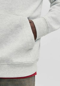 Vaaleanharmaa collegepaita resorilla varustetulla hihansuutimella, jossa on sivutasku. Kangas on hillityn tekstuuriinen, ja alla näkyy kerroksittainen punainen paita.