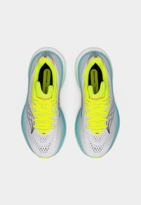 Ζεύγος παπουτσιών τρεξίματος Saucony με λευκά επάνω μέρη από δίχτυ, κορδόνια νέον κίτρινου χρώματος και τιρκουάζ ενδιάμεσα πέλματα, σε θέα από πάνω σε λευκό φόντο.