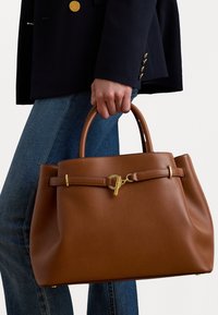 Személy nagy barna bőr kézitáskát tart, arany csattal, sötét zakót visel arany gombokkal és kék farmernadrággal.