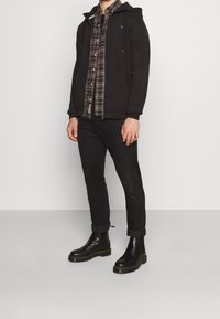 Mies, joka on pukeutunut mustaan vetoketjulliseen huppariin ruudullisen paidan päällä, mustiin ylös käärityihin housuihin ja mustiin nahkabootseihin, seisoo harmaata seinää vasten.