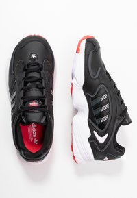 Black athletic shoes with gray stripes and red accents, featuring a textured upper, padded tongue, and a white and red rubber sole.