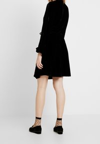 Vestido negro de terciopelo con mangas largas, cintura ceñida, falda acampanada y longitud a medio muslo. Combinado con zapatos planos negros y calcetines de red hasta el tobillo.