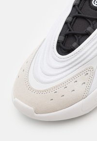 Zapatilla con una parte superior de malla blanca texturizada, una sección de tela negra, acentos de ante beige y detalles perforados en la puntera.