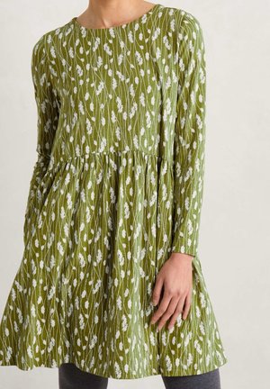 Kobieta ubrana w zieloną sukienkę z długim rękawem, w biały kwiatowy wzór, z marszczonym pasem, stojąca na jasnym, jednolitym tle.