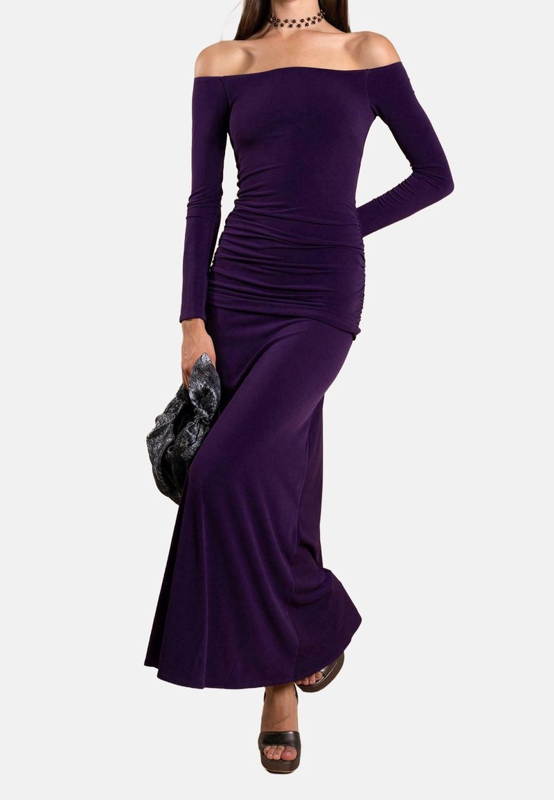 Femme portant une robe longue mauve ajustée à manches longues et épaules dénudées, tenant une pochette noire texturée et chaussant des talons ouverts noirs.