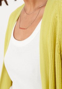 Pletený kardigan, živá limetkově zelená, s otevřeným předním dílem. Pod ním bílé žebrované tílko. Stříbrný řetízkový náhrdelník s růžovým akcentem.