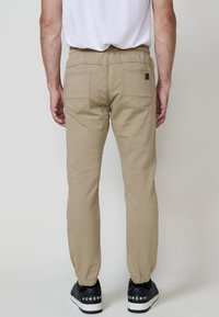 Pantaloni de drumeție din bumbac bej deschis, cu talie elastică, croi ajustat și două buzunare la spate. Purtați cu adidași negri cu accente albe.