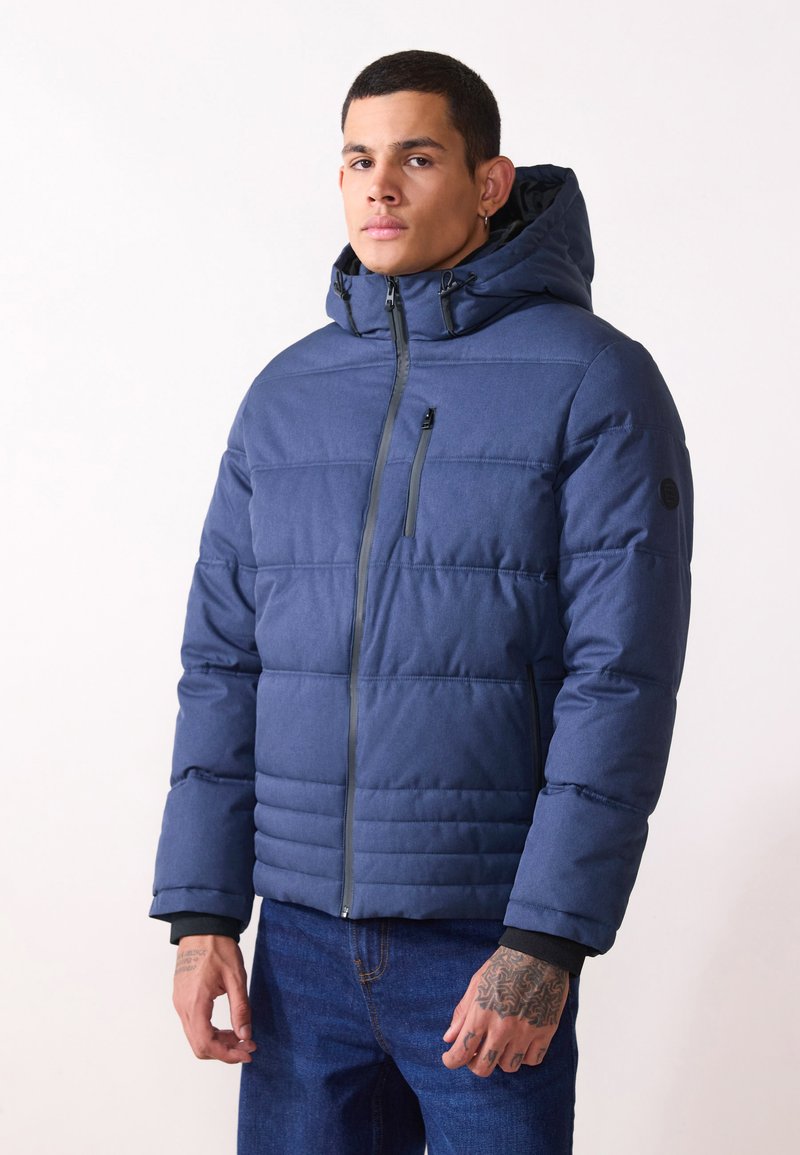 Chaqueta acolchada azul con capucha, que presenta un acolchado horizontal, cierre frontal con cremallera y un pequeño bolsillo en el pecho. Fabricada con un tejido de textura suave.