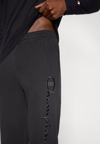 Μαύρο παντελόνι φόρμας Black Champion με ελαστική μέ ση, με ανάγλυφο μαύρο λογότυπο στο αριστερό πόδι. Το ύφασμα φαίνεται απαλό και άνετο.