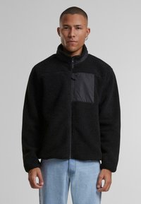 Jachetă din fleece negru cu guler înalt, fermoar frontal și buzunar neted negru în contrast. Suprafață texturată și moale, cu croială lejeră.