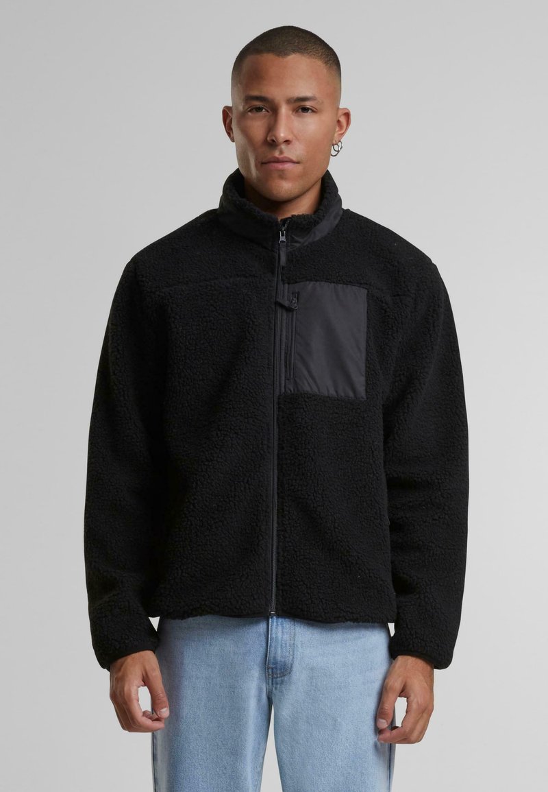 Jachetă din fleece negru cu guler înalt, fermoar frontal și buzunar neted negru în contrast. Suprafață texturată și moale, cu croială lejeră.