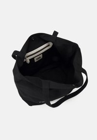 Černá plátěná taška přes rameno s dvěma popruhy, vybavená vnitřní kapsou na zip a krémovým zipem. Vnitřek je bez podšívky, což zdůrazňuje textilní strukturu.