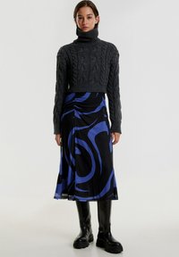 Grauer, kabelgestrickter, cropped Rollkragenpullover kombiniert mit einem schwarzen Rock, der blaue abstrakte Wirbel aufweist, getragen mit schwarzen Stiefeln.