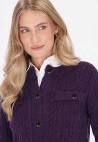 Fialový pletený cardigan s kabelovým vzorem, se胸ní kapsou a černými knoflíky, navrstvený přes bílou košili s límečkem.