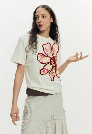 Jeune femme aux cheveux bouclés portant un t-shirt blanc avec un grand imprimé de fleurs rouges et roses et une jupe légère à volants, posant les mains levées.