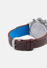 Bracelete de relógio de couro castanho com forro azul, fivela de metal prateada e costura contrastante, mostrando as costas de um relógio de pulso com caixa em aço inoxidável.
