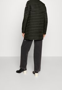 Mörkgrön puffjacka med horisontellt quiltad design, knälång, tillsammans med lösa svarta jeans och svarta klackskor.