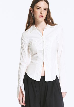 Jeune femme aux longs cheveux bruns portant un chemisier blanc cintré à manches longues et un pantalon noir ample, debout devant un fond blanc.