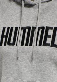 Sweat à capuche gris avec un logo "HUMMEL" brodé en noir. Comprend des cordons de serrage avec embouts gris et un tissu doux et texturé.