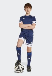 Marinfärgad fotbollsklädsel med en kortärmad tröja med vita ränder, matchande shorts och blå strumpor. Spelaren står med en fotboll.