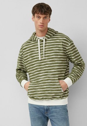 Młody mężczyzna z krótkimi brązowymi włosami, ubrany w zieloną i białą, paskowaną bluzę z kapturem i kieszenią z przodu oraz jasnoniebieskie dżinsy, stojący na jednolitym tle.