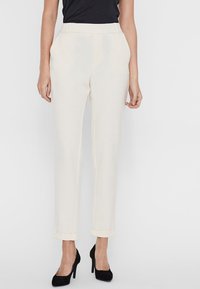 Pantaloni a tubo color crema con una trama liscia, caratterizzati da un risvolto e passanti per cintura, abbinati a tacchi alti neri.