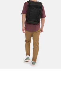 Mies, joka käyttää viininpunaista paitaa ja vaaleanruskeita housuja, joiden lahkeet on kierretty, kantaa suurta mustaa suorakulmaista reppua ja kävelee valkoista taustaa vasten.