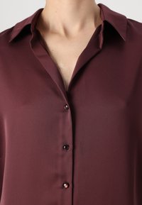 Bordová blúzka s klasickým golierom, so hladkou textúrou, zapínaním na gombíky vpredu a jemným leskom. Obsahuje dva gombíky na výstrihu.