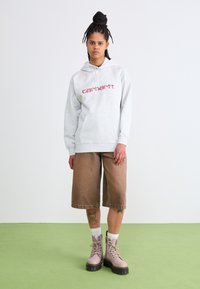 Felpa grigia con testo rosso, pantaloni corti in corduroy marrone e stivali rosa chiaro con lacci e suole spesse. Calzini bianchi visibili.