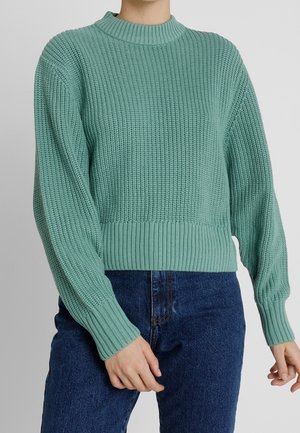 Maglione lavorato a maglia verde chiaro con texture a coste, collo alto e maniche lunghe, abbinato a jeans in denim blu scuro.