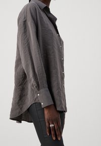Graues Langarm-Hemd mit Knopfleiste aus strukturierter Baumwolle, mit einer Brusttasche und silbernen Knöpfen. Kombiniert mit engen schwarzen Hosen.