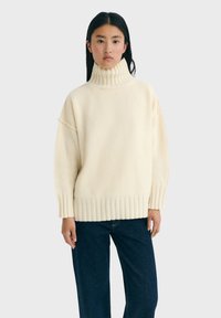 Pull en maille couleur crème avec un col haut côtelé, des épaules tombantes et un ourlet côtelé. Texture douce, coupe décontractée et design minimaliste.