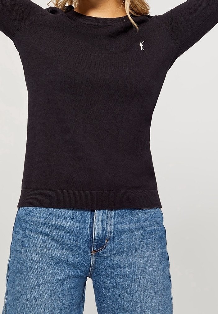 Femme portant un pull noir à manches longues avec une petite figure blanche brodée sur la poitrine et un jean en denim bleu clair.