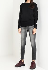 Maglione lavorato a maglia nera con accenti di perle sulle maniche, abbinato a jeans skinny grigi e sneakers bordeaux. Outfit casual con tessuti strutturati.