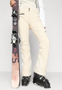 Μπεζ παντελόνια σκι με εφαρμογή που έχει ραφή διαθέτουν δύο μπροστινά κουμπιά και μία τσέπη με φερμουάρ. Μαύρα μποτάκια σκι με μεταλλικές αγκράφες. Σκι με σκούρο σχέδιο και δεσμά.