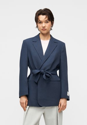 Tânăr cu păr scurt și închis la culoare, purtând un sacou bleumarin cu curea peste un top alb și pantaloni gri deschis.