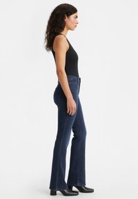 Zwarte mouwloze top en donkerblauwe flared jeans worden gedragen met zwarte enkellaarzen; de jeans hebben standaardstiksels en een vijf-pocket ontwerp.
