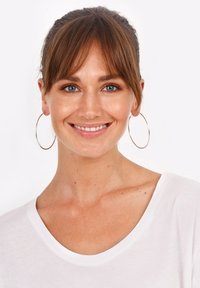 Pendientes de aro plateados complementan la cara sonriente de una modelo que lleva una camiseta blanca de escote redondo, con cabello castaño hasta los hombros y un maquillaje natural.