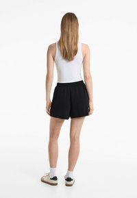 Zwarte shorts met een elastische tailleband, met een relaxte pasvorm. Draagt samen met een witte tanktop en casual schoenen, gezien vanaf de achterkant.