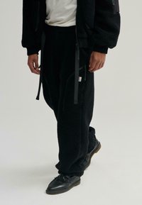 Czarne dresowe spodnie z luźnym krojem, z zamykanymi na zamek kieszeniami i sznurkiem w pasie. Noszone z czarnymi, frędzlami zdobionymi butami.