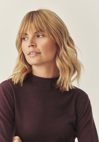 Hnědý svetr s dlouhým rukávem, mock neck, měkká textura a volný střih. Modelka má vlnité blond vlasy a přirozený make-up.