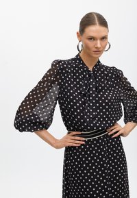 Svart blus med prickar, genomskinliga puffärmar, detalj med sjalknytnot vid halsen och elastisk midja. Mjuk textur och subtil lyster.