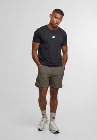 Mladý muž stojící s rukama v kapsách, oblečený v černém tričku, olivových šortkách, světlých béžových podkolenkách a bílých teniskách na jednobarevném pozadí.