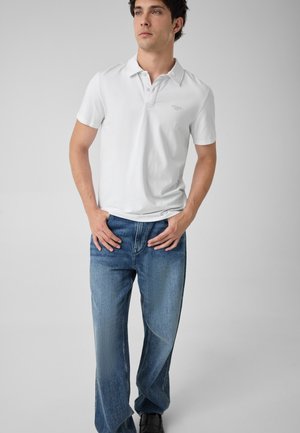 Jeune homme portant un polo blanc à manches courtes et un jean bleu, debout les mains près des poches devant un fond gris uni.