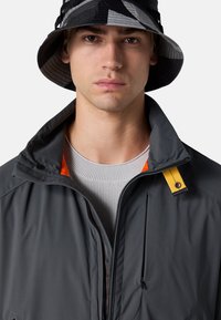 Grau Jacke mit hohem Kragen, orangefarbener Akzent am Hals und gelber Hardware. Getragen über einem hellgrauen strukturierten Pullover und einem schwarzen Bucket Hat.