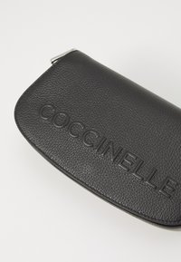 Clutch in pelle nera con superficie texture, con il marchio "COCCINELLE" embossato sulla parte anteriore e una forma liscia e curvata.