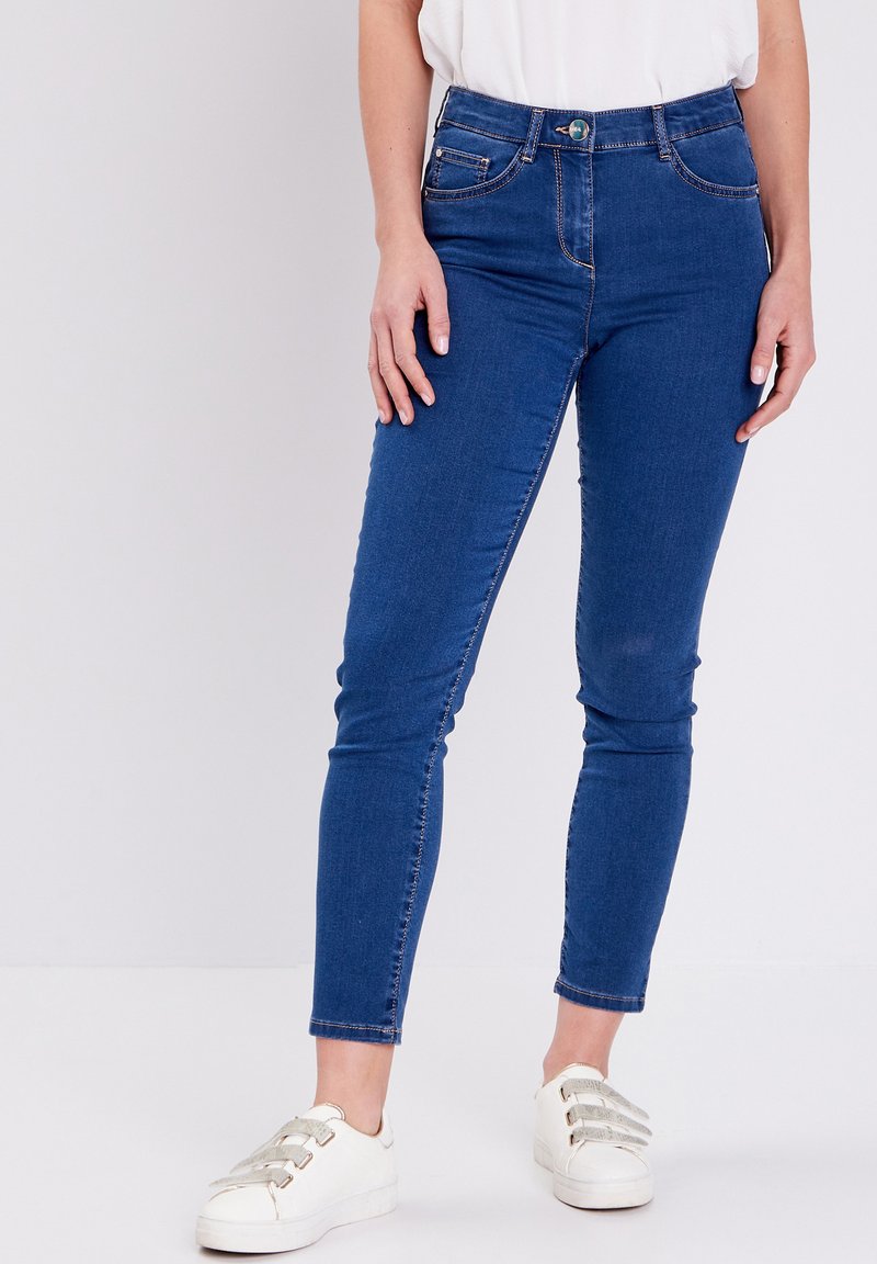 Blauwe denim skinny jeans met een medium hoge taille, vijf zakken en contrasterende stikdetails, gecombineerd met witte sneakers.