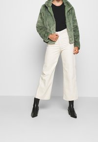 Giacca in pelliccia sintetica verde, top fitted nero, pantaloni a gamba larga color crema e stivali alla caviglia neri con punta quadrata. Texture morbida, vestibilità casual e stile a strati.